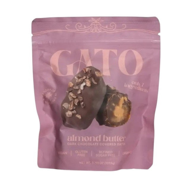A purple pouch of GATO DATES - CHOCOLATE COVERED ALMOND BUTTER DATES from GATO DATES displays almond butter dark chocolate covered Medjool dates, with text emphasizing that the product is vegan, plant-based, gluten-free, refined sugar-free, organic, and made with only 7 ingredients. The front of the pack features two chocolate-covered dates.