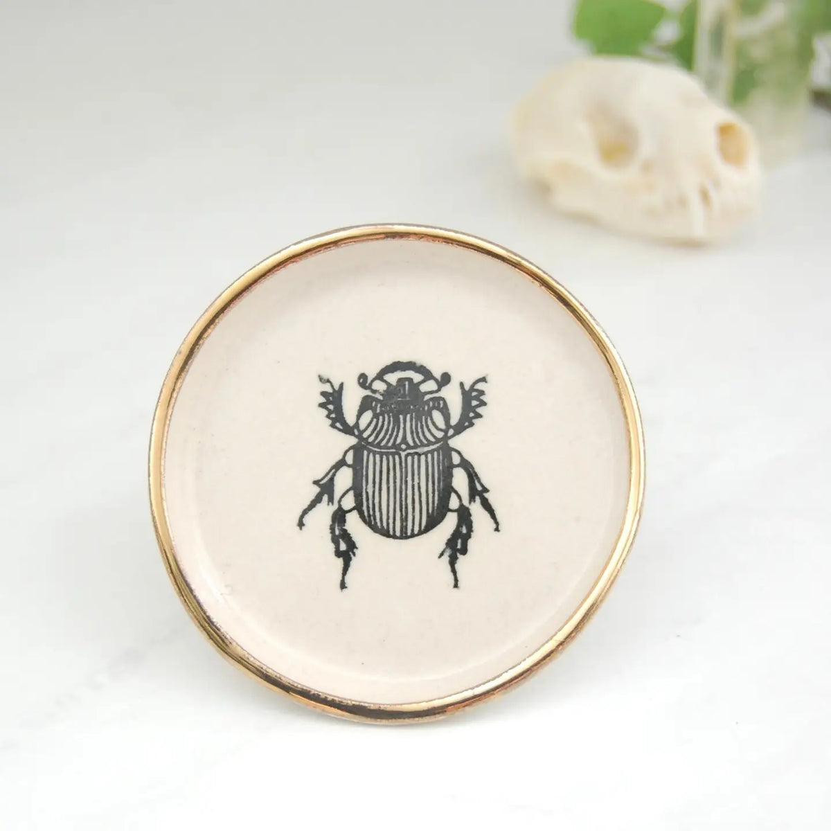 The COMPOST DESIGN - LITTLE BEETLE TINY DISH 3&quot;, with a black beetle illustration and 22k gold rim, is set on a white surface alongside a small skull-shaped object and a green plant.