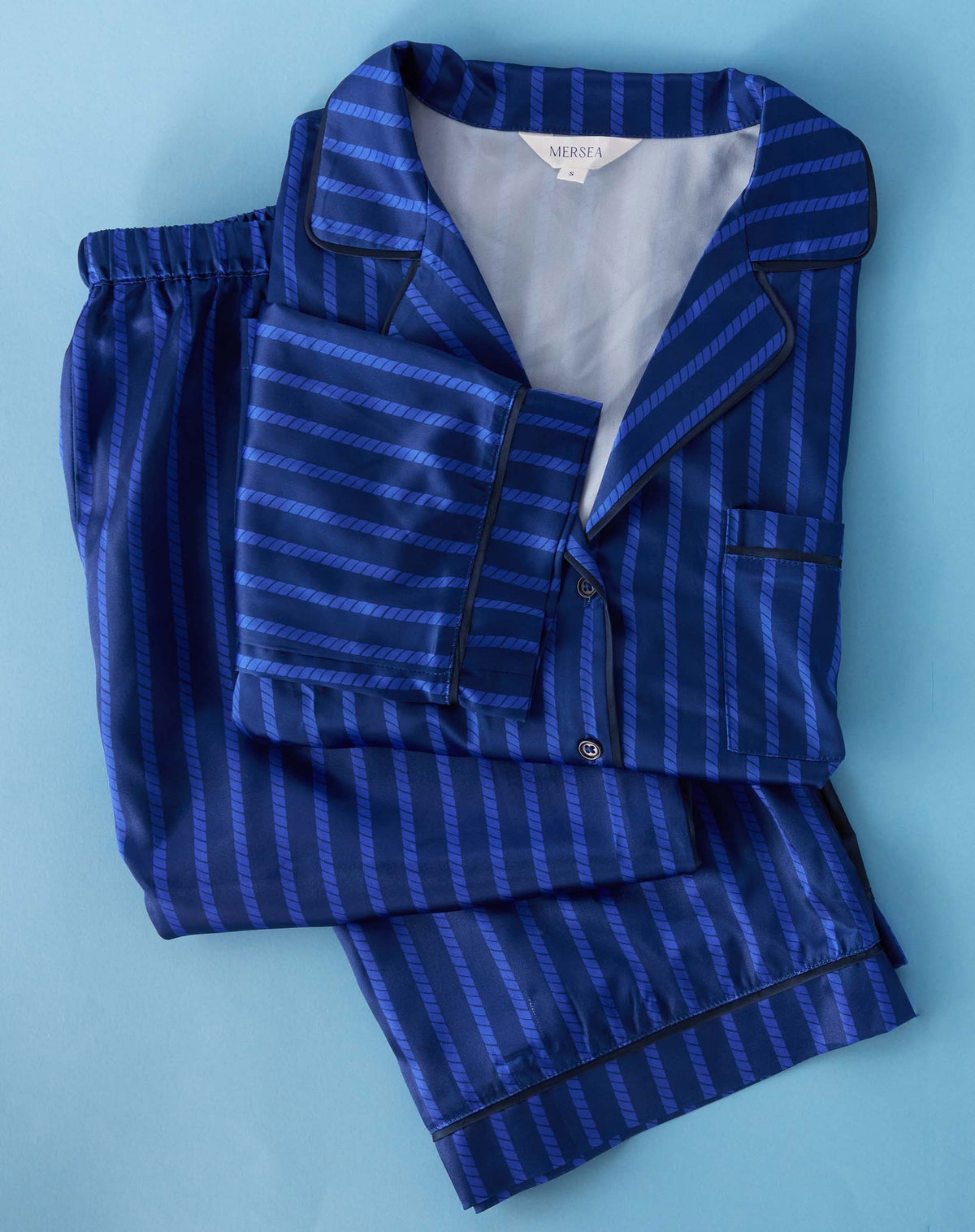 A person stands barefoot indoors, wearing the MERSEA Carousel Stripe Satin Sailor Pajama Set in Blue. They are leaning on a light gray chair, with a tripod lamp and large windows in the background, perfectly capturing the elegance of luxury sleepwear from MERSEA.