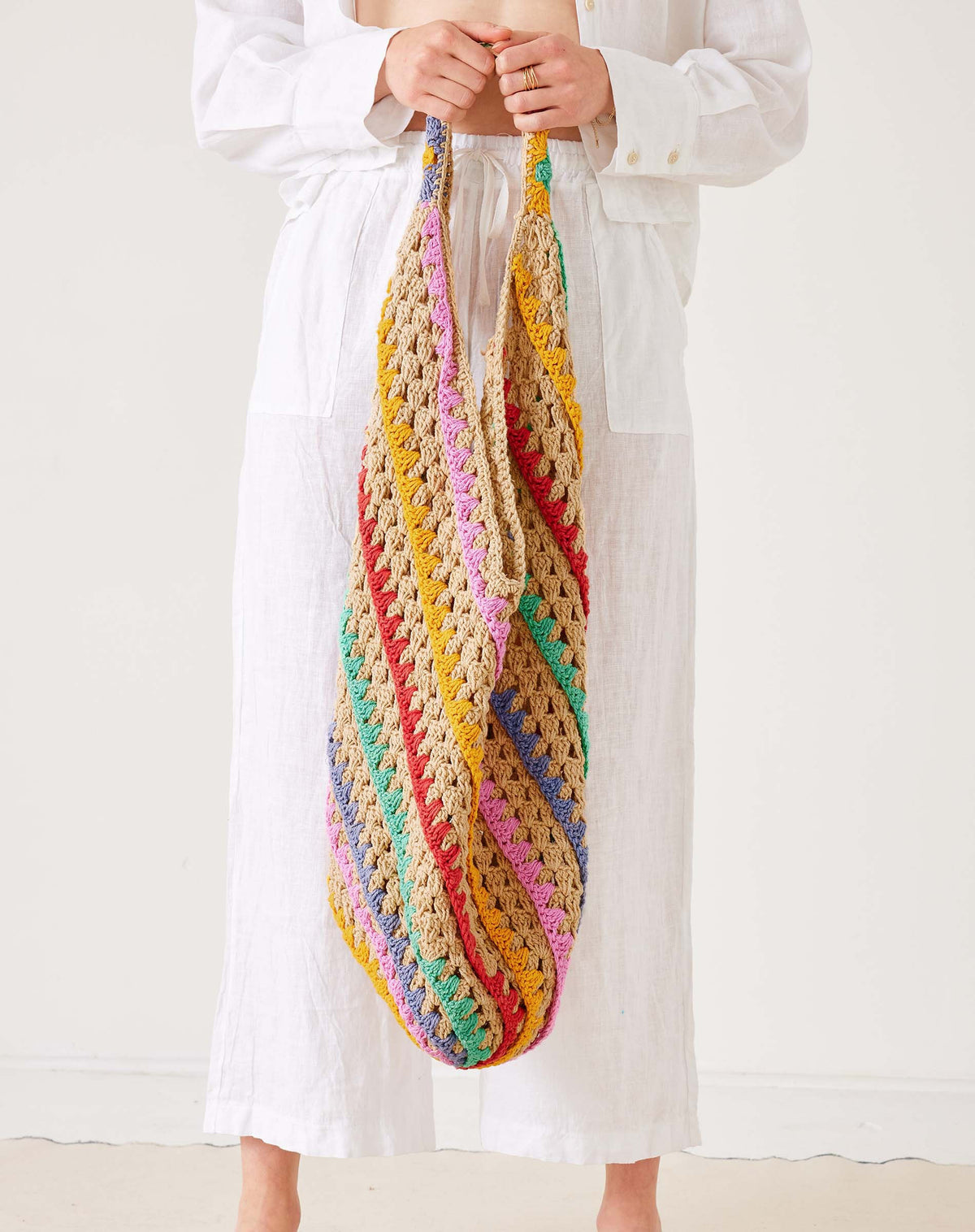 A person in white holds the MERSEA - CARLY CROCHET BOHO BAG by MERSEA, featuring large, colorful diagonal stripes of yellow, pink, red, green, and purple against a plain light background.