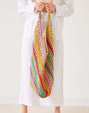 A person in white holds the MERSEA - CARLY CROCHET BOHO BAG by MERSEA, featuring large, colorful diagonal stripes of yellow, pink, red, green, and purple against a plain light background.