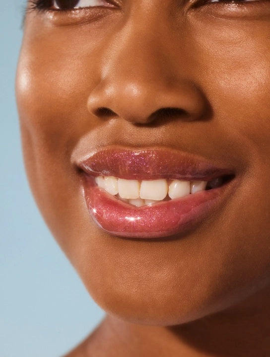 A close-up of a person smiling with glossy pink lips—enhanced by LANOLIPS - LIQUID LANOLIN LIP WATER from LANOLIPS—and smooth brown skin against a light blue background.