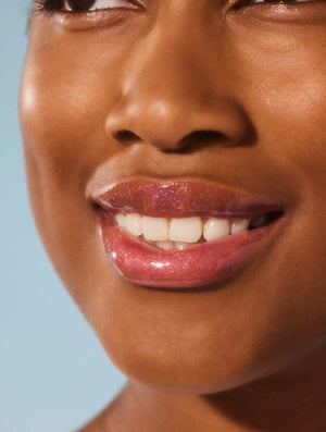 A close-up of a person smiling with glossy pink lips—enhanced by LANOLIPS - LIQUID LANOLIN LIP WATER from LANOLIPS—and smooth brown skin against a light blue background.