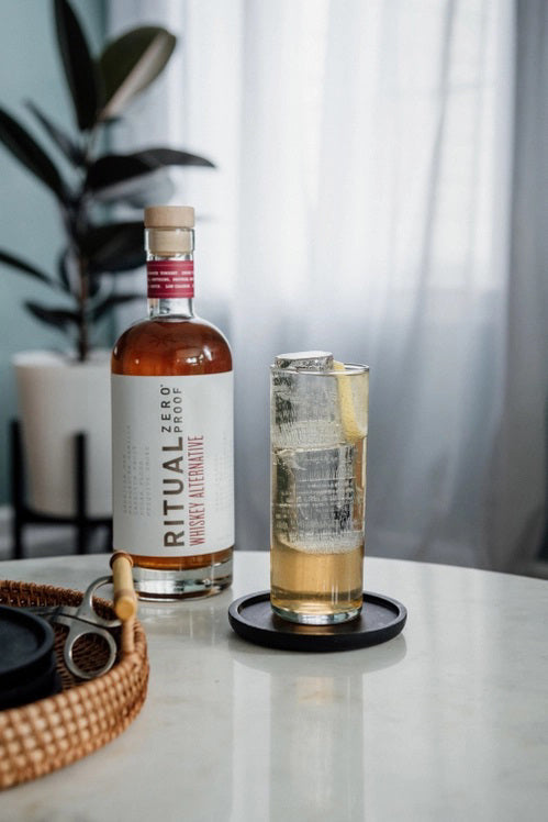 A bottle of RITUAL ZERO PROOF-WHISKEY ALTERNATIVE caters to health-conscious consumption. The bottle has a white label with &quot;RITUAL ZERO PROOF&quot; in black text and &quot;Whiskey Alternative&quot; in red text. The cap is topped with a red seal, and the alcohol-free whiskey inside is amber-colored.