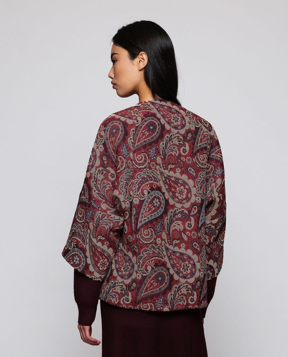 A woman stands against a plain background wearing the MIRTO 1956 Burgundy Paisley Jacquard Jacket—an oversized, cotton-blend piece in rich red and purple tones from MIRTO 1956—styled over a long burgundy dress and black shoes. The relaxed fit of the jacket complements her straight dark hair and neutral expression.