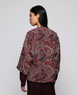 A woman stands against a plain background wearing the MIRTO 1956 Burgundy Paisley Jacquard Jacket—an oversized, cotton-blend piece in rich red and purple tones from MIRTO 1956—styled over a long burgundy dress and black shoes. The relaxed fit of the jacket complements her straight dark hair and neutral expression.