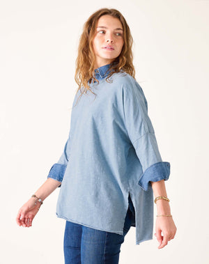 A woman with wavy light brown hair wears the MERSEA - CATALINA SLUB TEE IN SHORE BLUE STITCHES over a collared shirt and dark jeans, standing against a plain background and looking to her left.