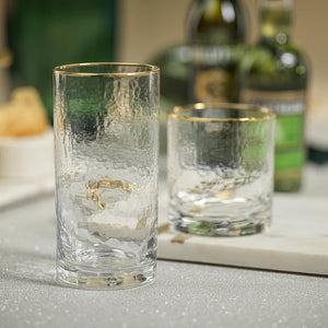 A clear, cylindrical NEGRONI HAMMERED DOUBLE OLD FASHIONED GLASS by ZODAX with a slightly textured surface and a thin gold rim at the top, boasting a capacity of 347 ml. The glass is empty and positioned against a plain white background.