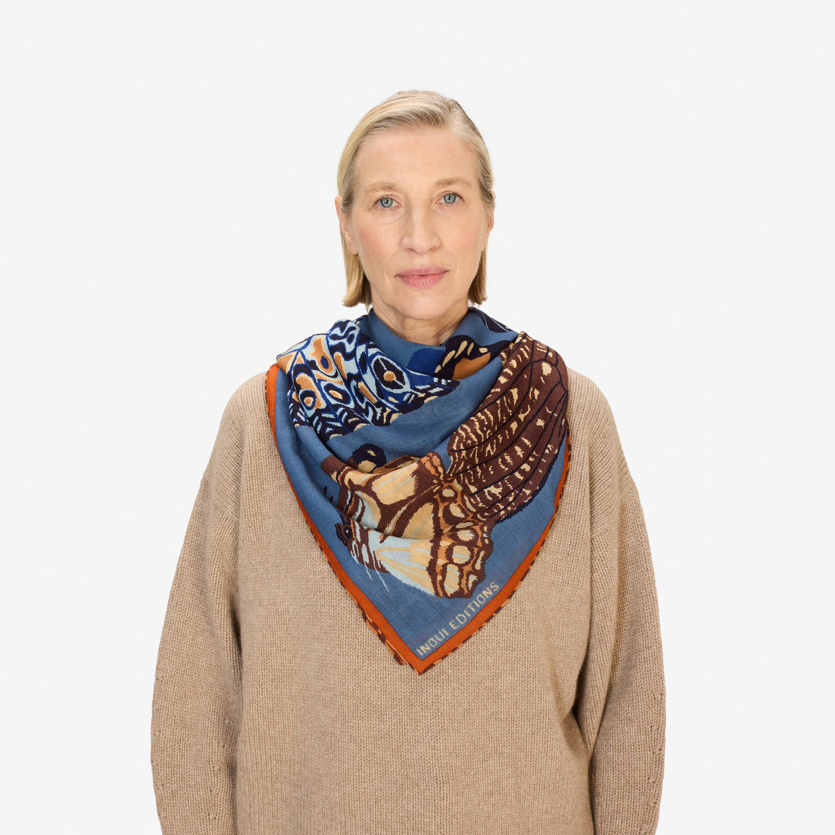 A woman wears the INOUI EDITIONS PAPILLON (BUTTERFLIES) wool square scarf in duck blue, featuring abstract nature-inspired designs, paired with a beige sweater. Only her lower face and neck are visible.