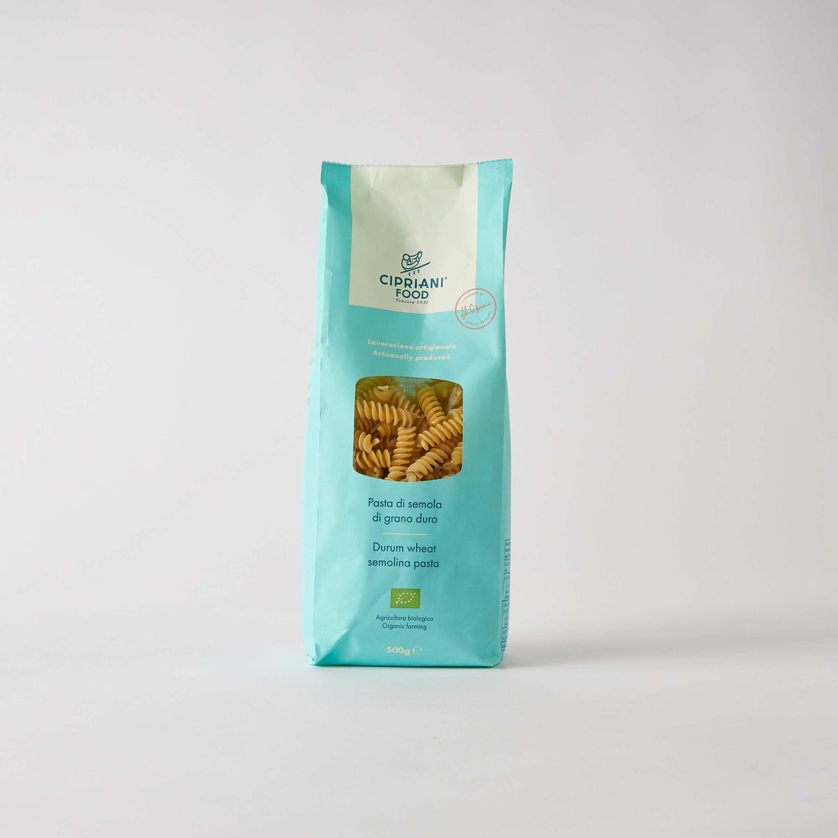 A turquoise bag of Atalanta Corporation's Cipriani Organic Durum Wheat Fusilli, 500g, features a clear window displaying spiral-shaped pasta made from Italian organic semolina. The bag has a white label on top and text in both Italian and English, set against a plain white background.