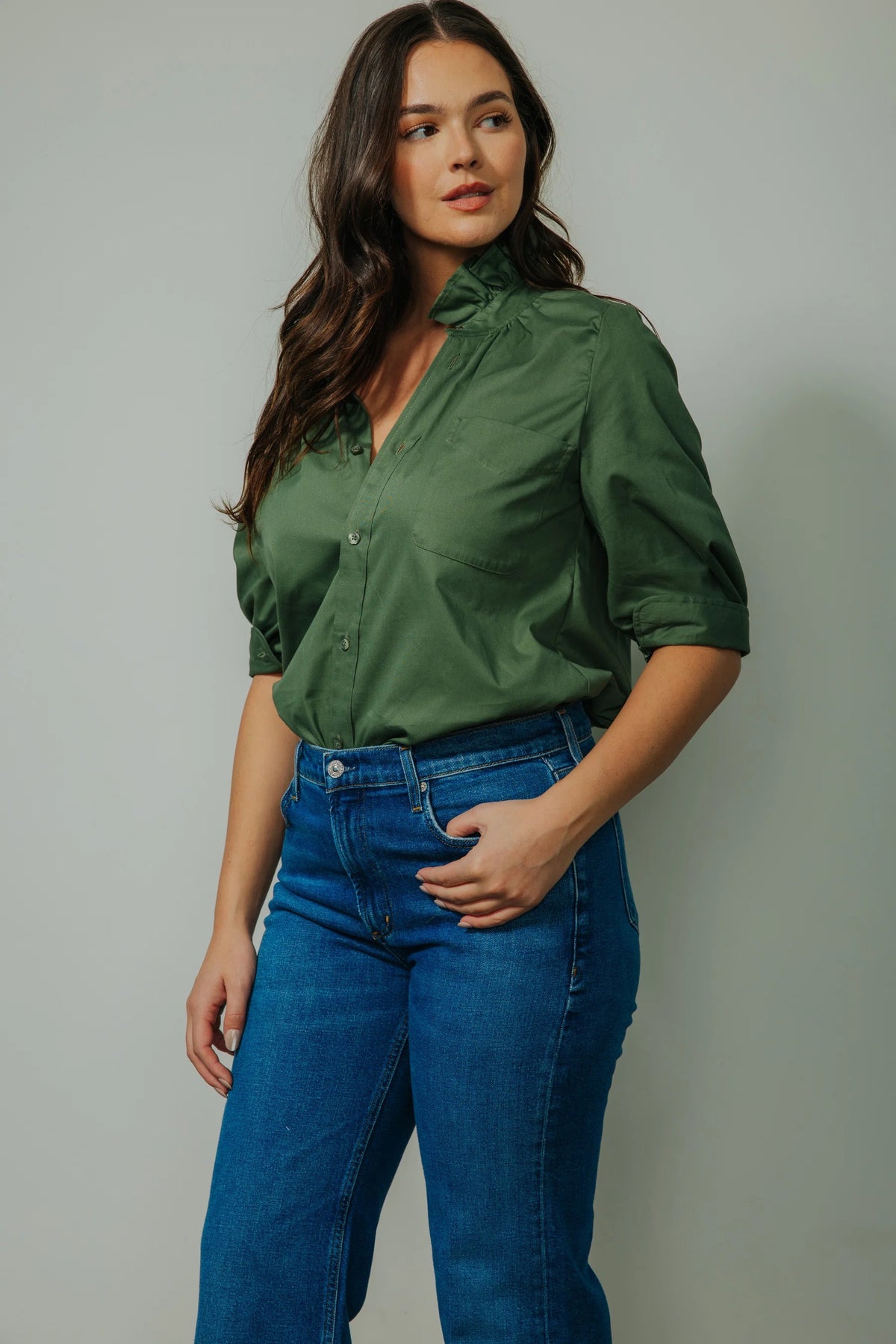 A woman in the KMJ - Southern Lover Shirt with a ruffle collar and blue jeans, featuring the iconic KMJ tuck, stands confidently beside a horse, holding its reins amidst green foliage. She's elegantly posed with jewelry, embodying Southern-inspired charm.