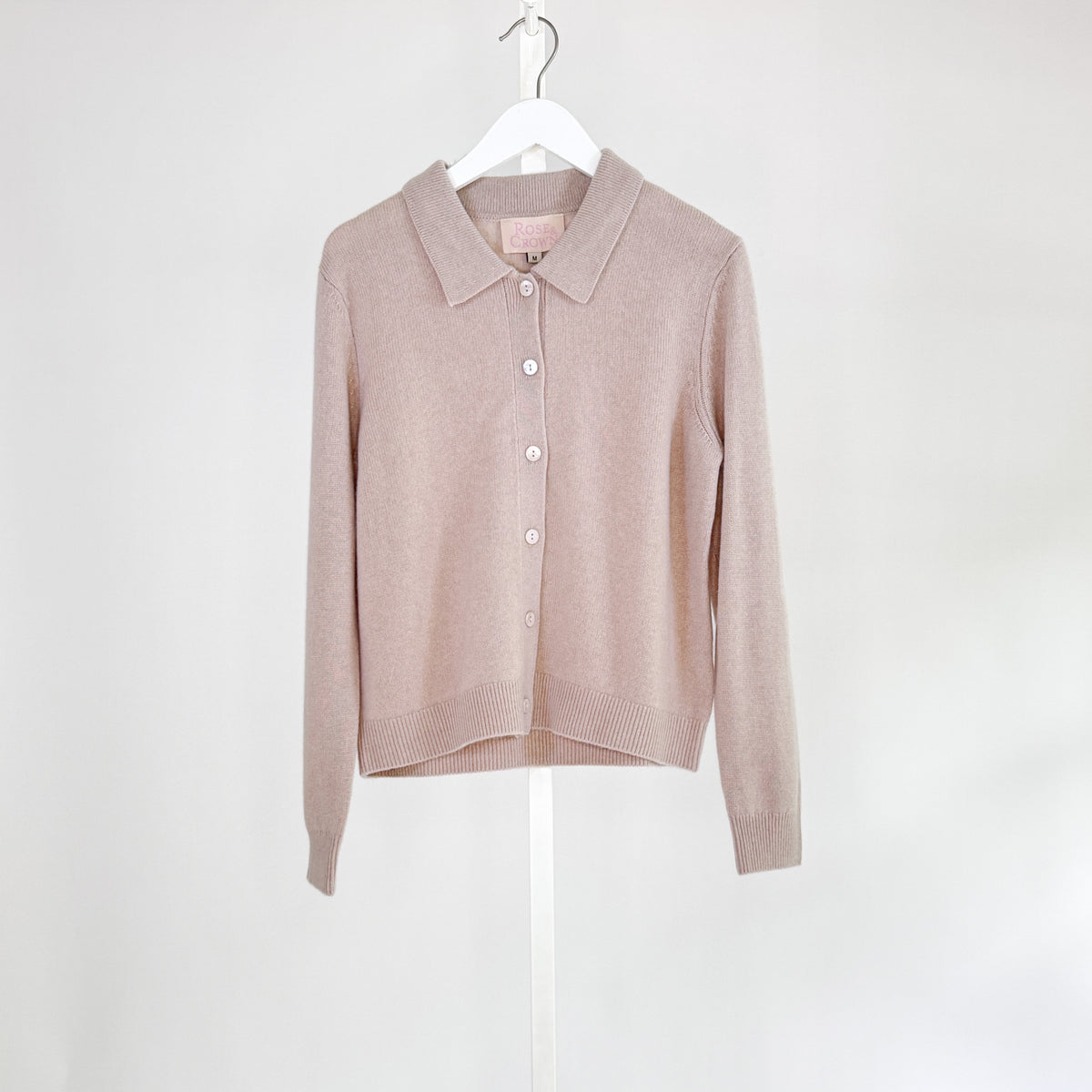 The ROSE &amp; CROWN - CASHMERE CARDIGAN, a luxury long-sleeve button-up in custom-dyed magenta, hangs on a white hanger against a light gray background.
