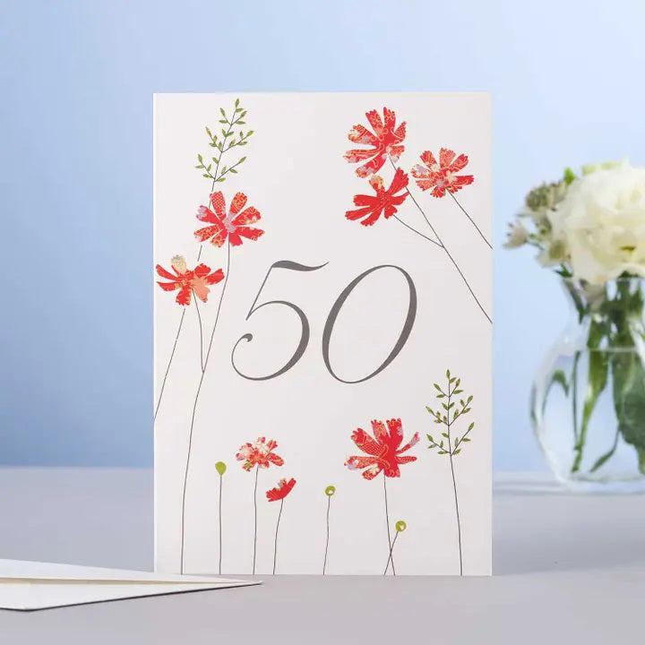 The ELOISE HALL - COSMOS 50TH BIRTHDAY CARD by ELOISE HALL LTD features the number "50" in elegant script, surrounded by red Cosmos flowers with green stems and leaves. The card stands on a light blue surface, while a blurred glass vase containing white flowers and greenery in the background accentuates the beauty of this serene scene.