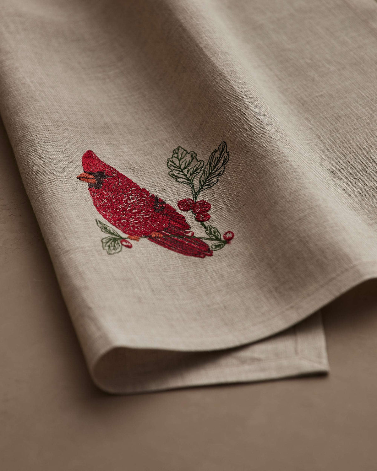 A RED CARDINAL LINEN TEA TOWEL by CORAL & TUSK made of beige natural flax fiber, embroidered with a red cardinal perched on a branch with berries and green leaves.