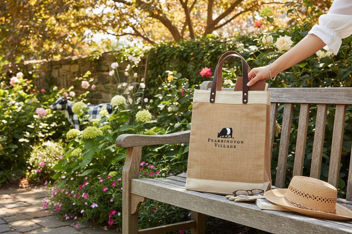 The FEARRINGTON LIFESTYLE COLLECTION - BELTIE JUTE TOTE by STRAN PROMOTIONAL SOLUTIONS is a stylish, eco-friendly tan and beige jute bag with dark leather handles and an embroidered black and white badger above "Fearrington Village.