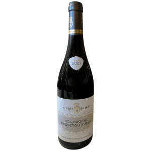 A bottle of ALBERT BICHOT ‘PASSETOUTGRAIN’ 2021 red wine stands on a white table in front of a wine rack.