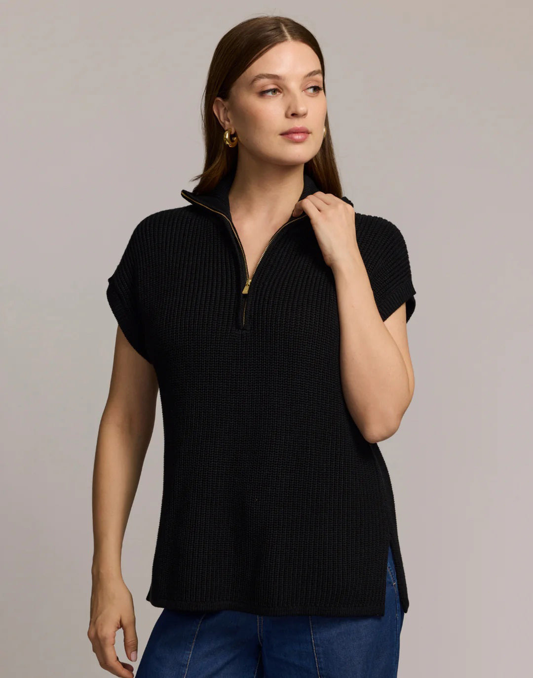 A woman with straight brown hair wears the HINSON WU - BROOKE SLEEVELESS SWEATER VEST in black with a gold-tone half-zip and blue jeans, standing facing forward against a plain light grey background.