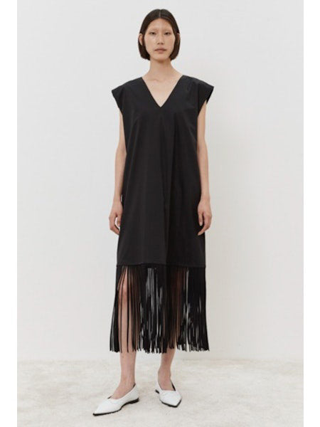 The person models the MEIMEIJ - FRINGE DRESS, a cotton, sleeveless black dress with a V-neckline and knee-length flowing fringes. They pair it elegantly with white shoes against a plain white background.