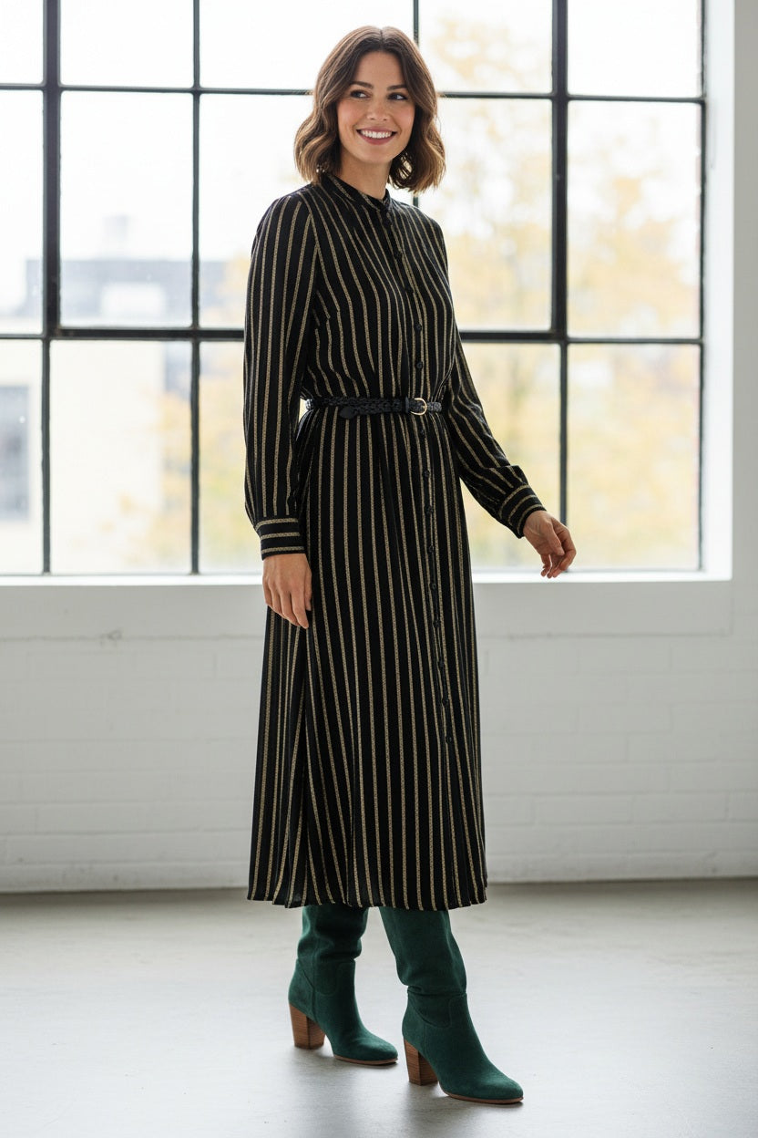 A woman in the MOMONI Aubert Dress in Lurex Gauze stands by a woven chair, gazing down as her hair partially covers her face.