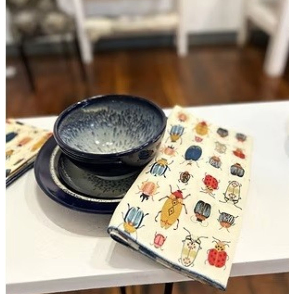 Introducing the DOT AND ARMY - BEETLE DINNER NAPKIN by DOT AND ARMY: a folded white cotton napkin adorned with a vibrant pattern of beetles and bugs in red, blue, yellow, and black hues, finished with blue edge stitching. Perfect as reusable napkins or charming beetle dinner accents for your table setting.