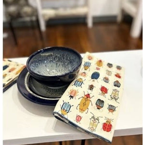Introducing the DOT AND ARMY - BEETLE DINNER NAPKIN by DOT AND ARMY: a folded white cotton napkin adorned with a vibrant pattern of beetles and bugs in red, blue, yellow, and black hues, finished with blue edge stitching. Perfect as reusable napkins or charming beetle dinner accents for your table setting.
