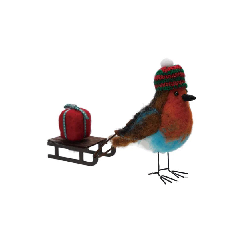 The CREATIVE COOP FELT ROBIN PULLING SLEIGH features a handmade wool felt robin in a red and green knit hat beside a sled with a red gift—perfect as festive holiday decor or a unique gift.