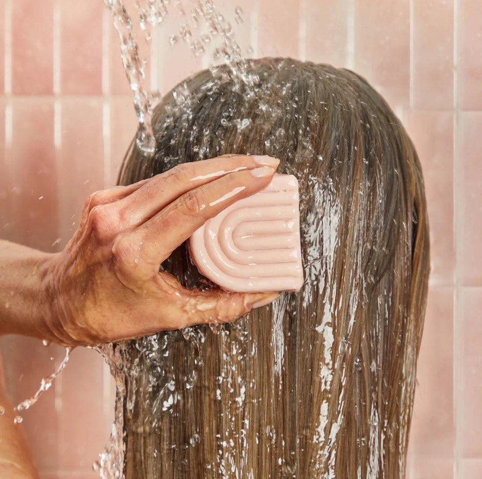 A pink KITSCH - CASTOR OIL NOURISHING CONDITIONER BAR and its box are shown with castor beans. The box notes it's color safe, pH balanced, vegan, and designed to nourish dry hair while controlling frizz.