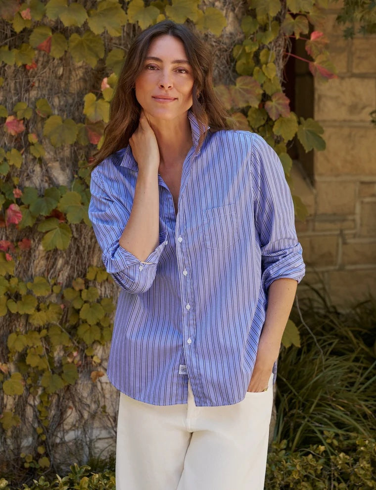 A woman stands in a sunlit room, smiling softly with wavy brown hair and one hand tucked into her pocket near a large window. She wears light blue jeans, tan loafers, and the FRANK & EILEEN EILEEN Relaxed Button Up Shirt in Italian Superluxe Cotton Multi Blue Stripe by FRANK & EILEEN. The shirt’s bust-flattering button placement adds a subtle touch of elegance to her effortlessly chic look.