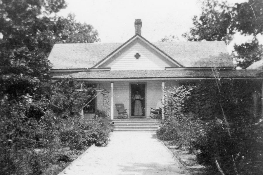 Black and white photo of a single-story house with a porch, two rocking chairs, and a person standing in the doorway. Lush plants and trees surround the walkway leading to the entrance.