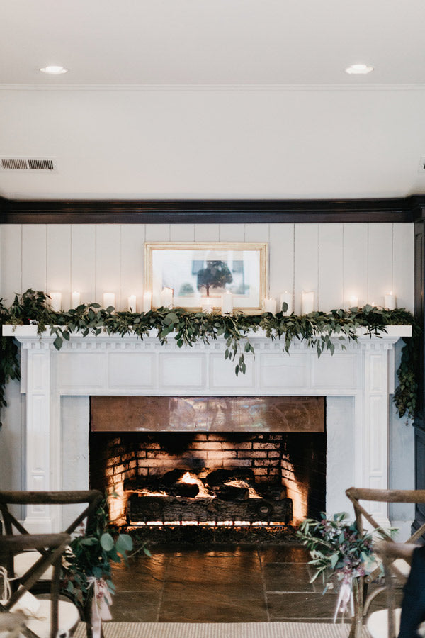 A white fireplace decorated with greenery and lit candles on the mantel. A framed picture hangs above the mantel, and logs are burning in the fireplace, creating a warm and cozy atmosphere.