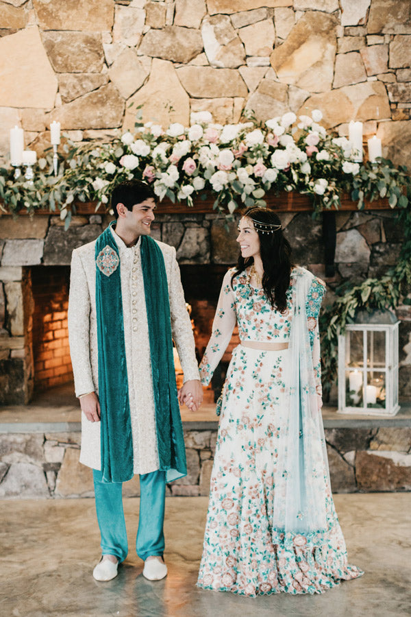 A couple in traditional South Asian attire stands holding hands in front of a stone fireplace decorated with flowers, greenery, and candles. The woman wears a floral lehenga; the man wears a cream sherwani with teal pants and scarf.