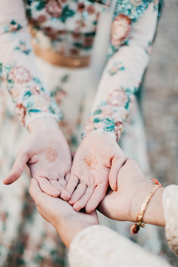 Two people in floral-patterned clothing hold out their hands, palms up, showing intricate henna designs. Another person gently cups their hands underneath, wearing a delicate bracelet.
