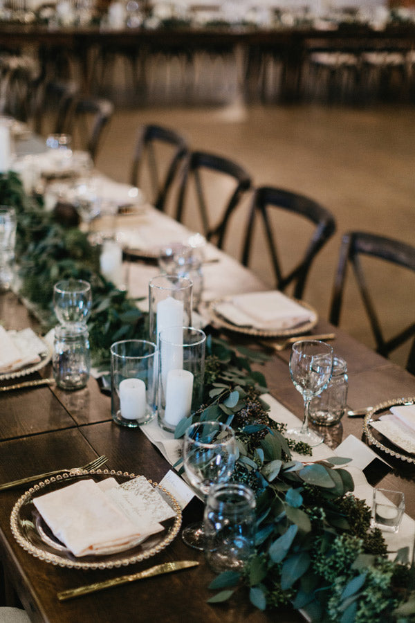 A long wooden table set for an event with elegant place settings, wine glasses, white napkins, gold-rimmed plates, greenery garlands, and glass candle holders with white candles.