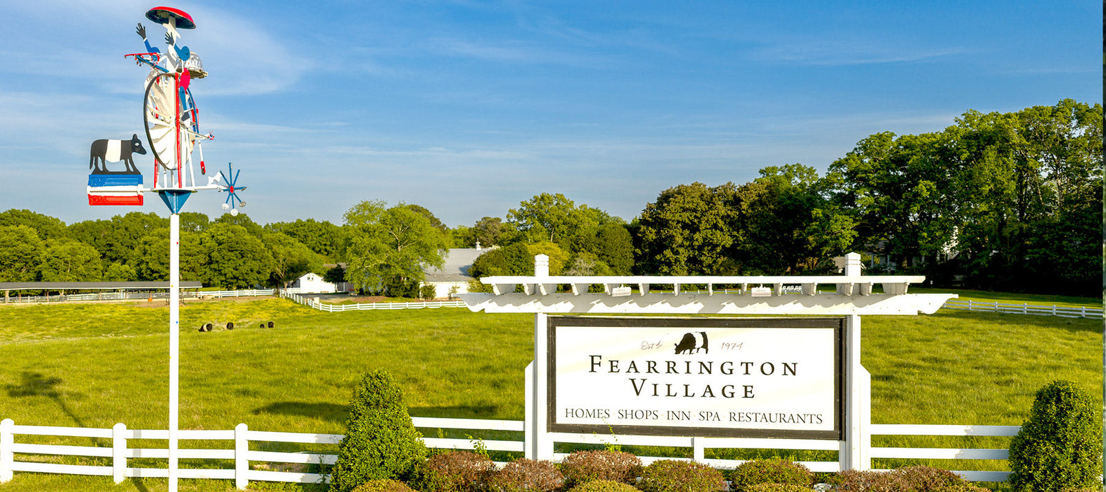 A white entrance sign for Fearrington Village stands near trimmed bushes and a white fence, with a colorful cow-themed weather vane and green trees in the background under a blue sky.