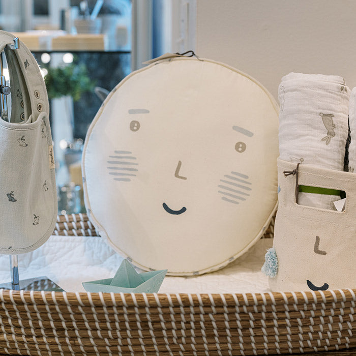 A round, cream-colored cushion with a simple smiling face and blush marks sits in a wicker basket next to soft fabric items, including a rolled-up blanket and small pouches, in a cozy, well-lit room.