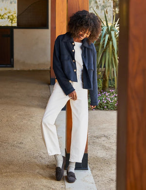 A woman with long brown hair wearing the FRANK & EILEEN Rowan Cropped Sweatshirt Utility Jacket in Triple Terry Classic Navy, paired with white pants, stands sideways and smiles outside near a white building with windows.