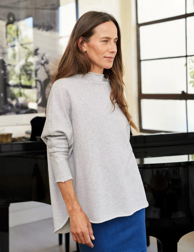 A woman with long brown hair stands smiling near a black grand piano in a modern, well-lit room. She wears the FRANK AND EILEEN - EFFIE LONG SLEEVE FUNNEL NECK CAPELET from FRANK &amp; EILEEN paired with blue jeans. A large window and a lamp are in the background, enhancing the cozy winter warmth of her stylish ensemble.