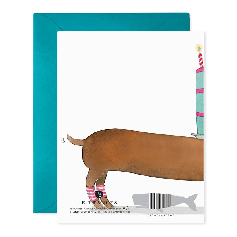 The E. FRANCES PAPER - SLINKY DOG BIRTHDAY GREETING CARD features a whimsical watercolor of a dachshund in pink and white socks carrying a cake with "Happy Birthday to You" on its back, complemented by a charming blue envelope background.