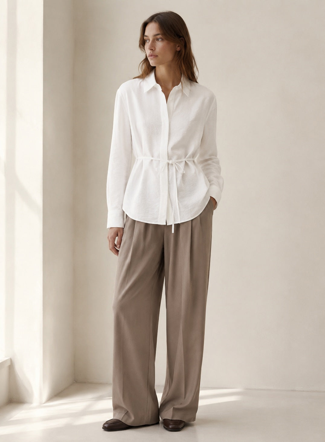 The ELEH LONG SLEEVE SHIRT WITH TIES by ELEH, crafted from a white Italian cotton blend, hangs neatly against a plain white wall. Featuring a collar and waist tie detail, this modern button-down silhouette is perfect for any occasion.