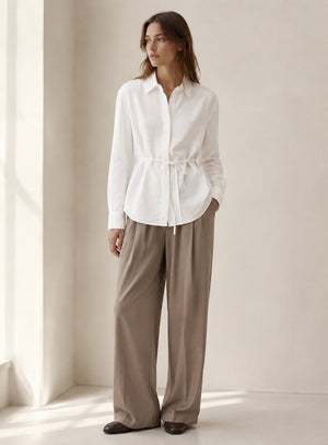 The ELEH LONG SLEEVE SHIRT WITH TIES by ELEH, crafted from a white Italian cotton blend, hangs neatly against a plain white wall. Featuring a collar and waist tie detail, this modern button-down silhouette is perfect for any occasion.