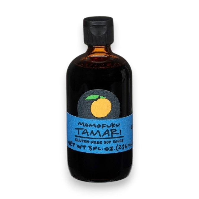 A dark glass bottle with a black cap and blue label reading "MOMOFUKU - TAMARI"—a premium, gluten-free soy sauce by MOMOFUKU—features an orange fruit graphic above the text. The bottle stands on a white surface.