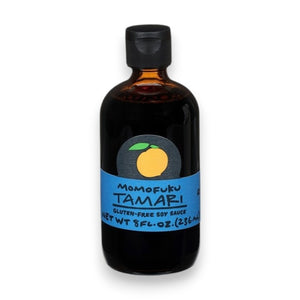A dark glass bottle with a black cap and blue label reading "MOMOFUKU - TAMARI"—a premium, gluten-free soy sauce by MOMOFUKU—features an orange fruit graphic above the text. The bottle stands on a white surface.