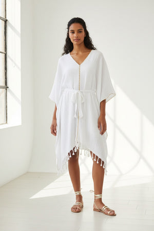 A woman with wavy hair smiles in the MERSEA - BEACH KAFTAN, a loose white Turkish cotton cover-up with fringe and gold trim, tied at the waist. She pairs it with sandals, showcasing MERSEA’s effortless resort style against a white background.