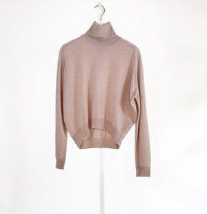 A woman with long brown hair smiles against a white background, wearing a ROSE & CROWN - CASHMERE TURTLENECK and wide-legged green pants, accessorized with necklaces, bracelets, and a large black handbag.