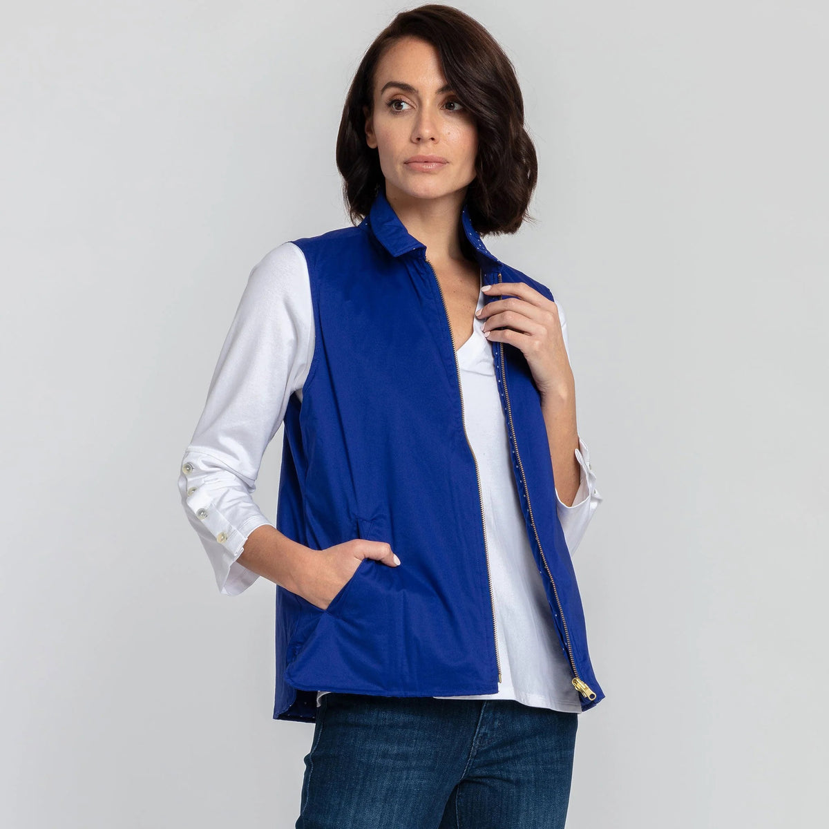 A woman with short brown hair is wearing the HINSON WU - LAUREN REVERSIBLE VEST in bright blue, paired with a white, long-sleeve blouse that features buttons on the cuffs. She completes her outfit with blue jeans and stands against a plain white background.