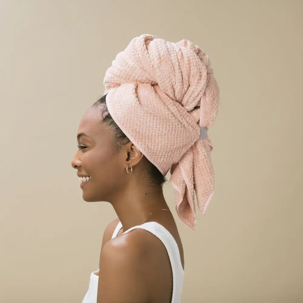 A person with short dark hair, wearing a white shirt, wraps their hair in the VOLO BEAUTY - HERO HAIR TOWEL LUXE and adjusts it with both hands in a bright, airy space filled with soft natural light.
