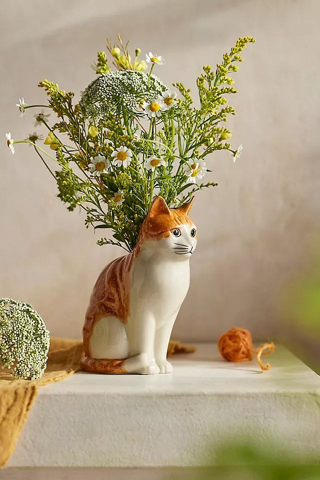 The QUAIL CERAMICS - SQUASH CAT FLOWER VASE by QUAIL DESIGNS is a hand-painted stoneware vase shaped like a sitting orange cat, with green foliage and yellow flowers appearing to grow from its back.