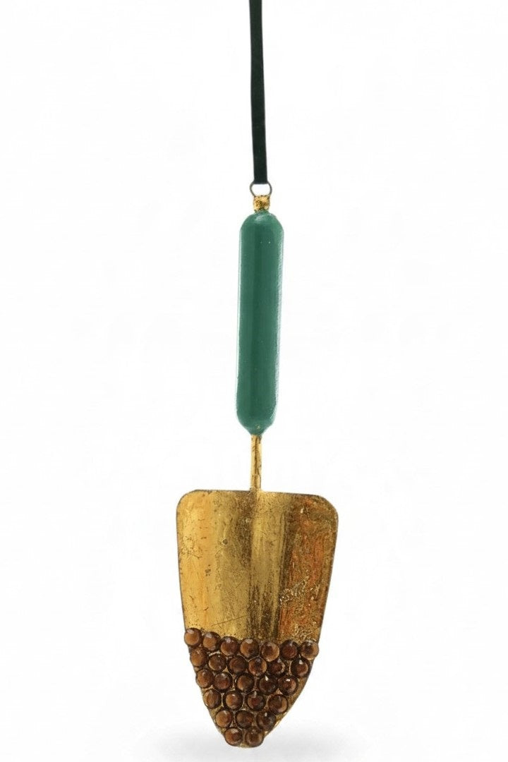 The JEWELED TROWEL ORNAMENT by Cody Foster is a unique holiday decoration shaped like a hanging garden trowel, featuring a gold-toned blade accented with brown crystals and a green handle.