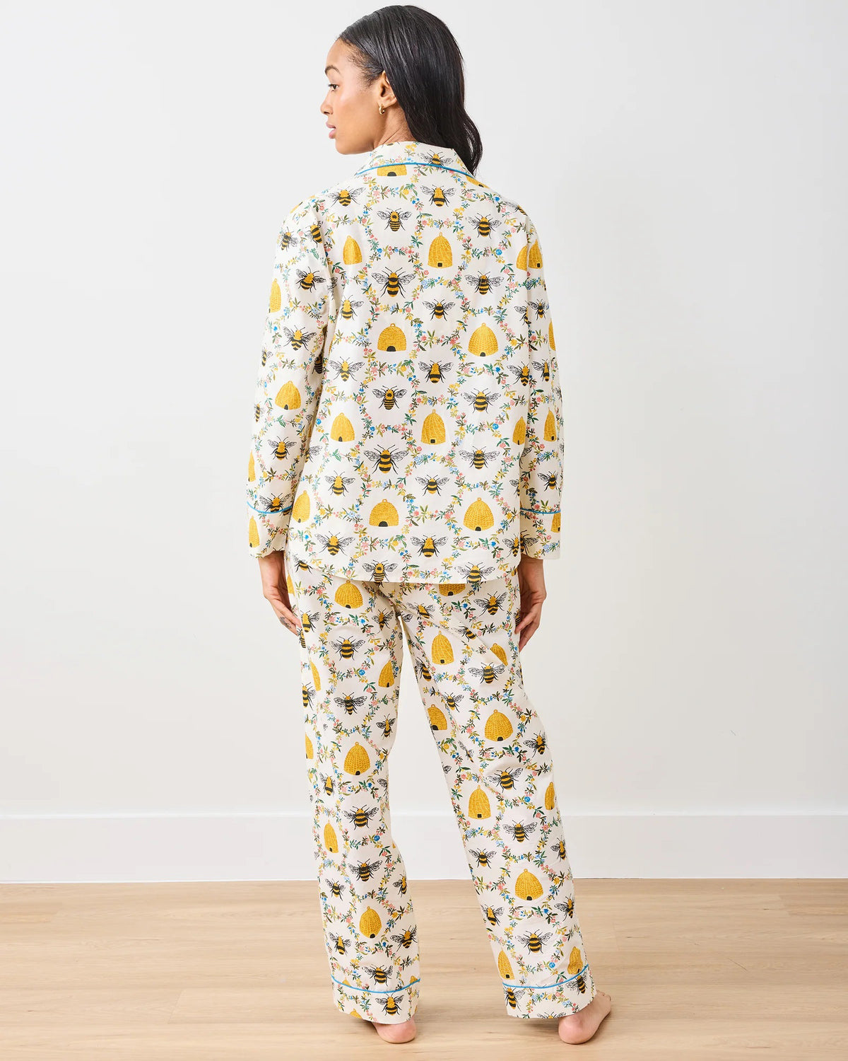 A person stands barefoot on a wooden floor wearing the PRINTFRESH - SWEET BEE COTTON PAJAMA SET. This organic cotton pajama is white with yellow sun and cloud patterns, accented by black piping, resembling bee pajamas against a plain white wall.