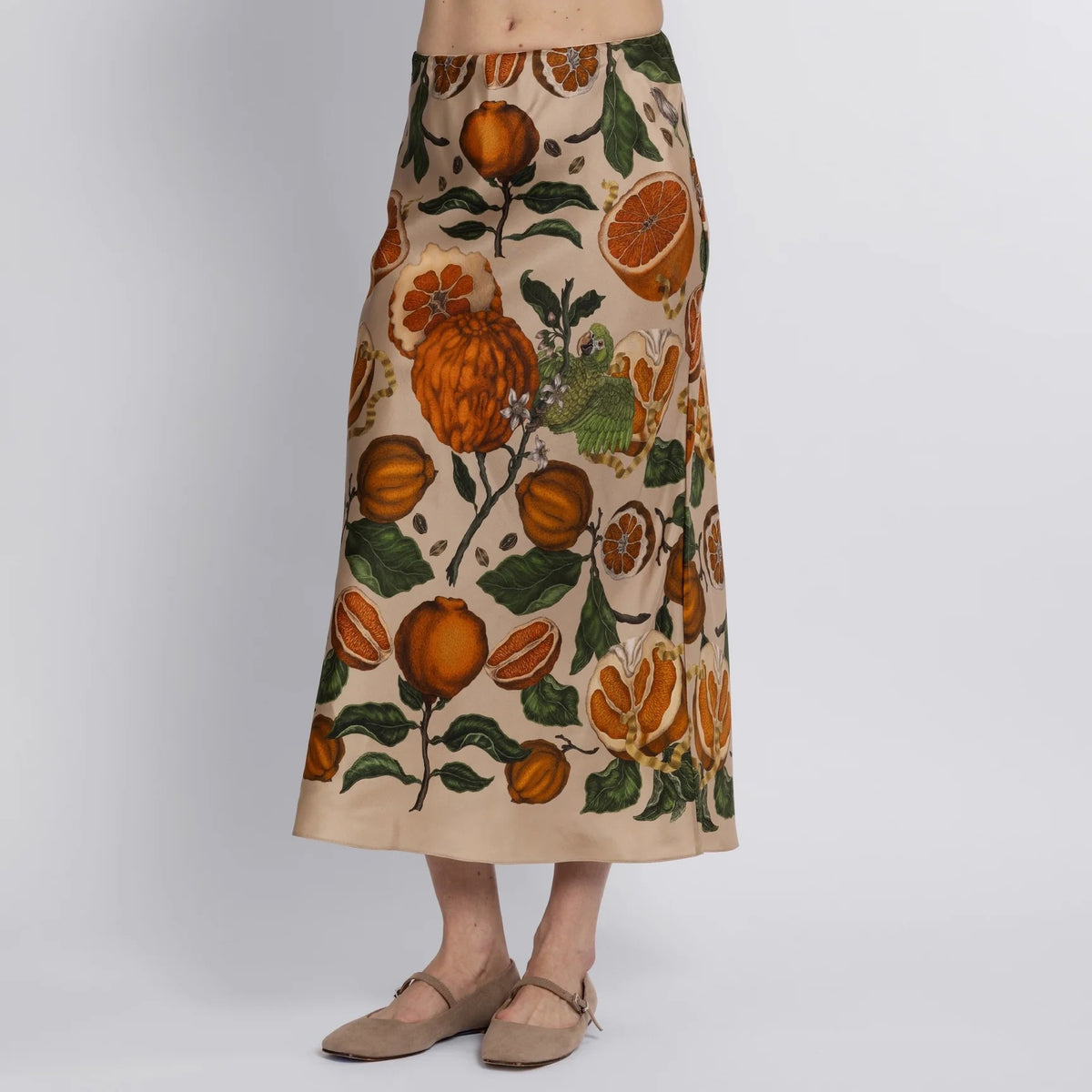 A person wears a SABINA SAVAGE long beige skirt, featuring an exotic print of vibrant florals, oranges, and leaves. Crafted from fine silk and draping elegantly to their ankles, they&#39;re barefoot against a plain white backdrop. Made by SABINA SAVAGE ENGLAND.